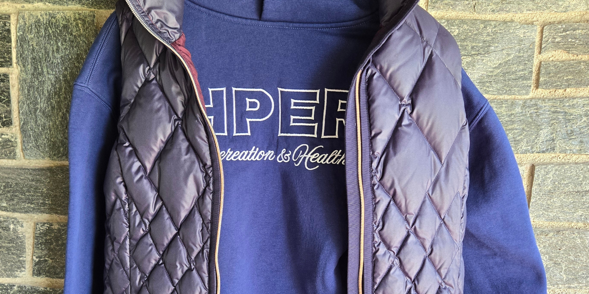 Image of a cobalt blue hoodie with a blue puffer vest hanging in front of a gray brick background.