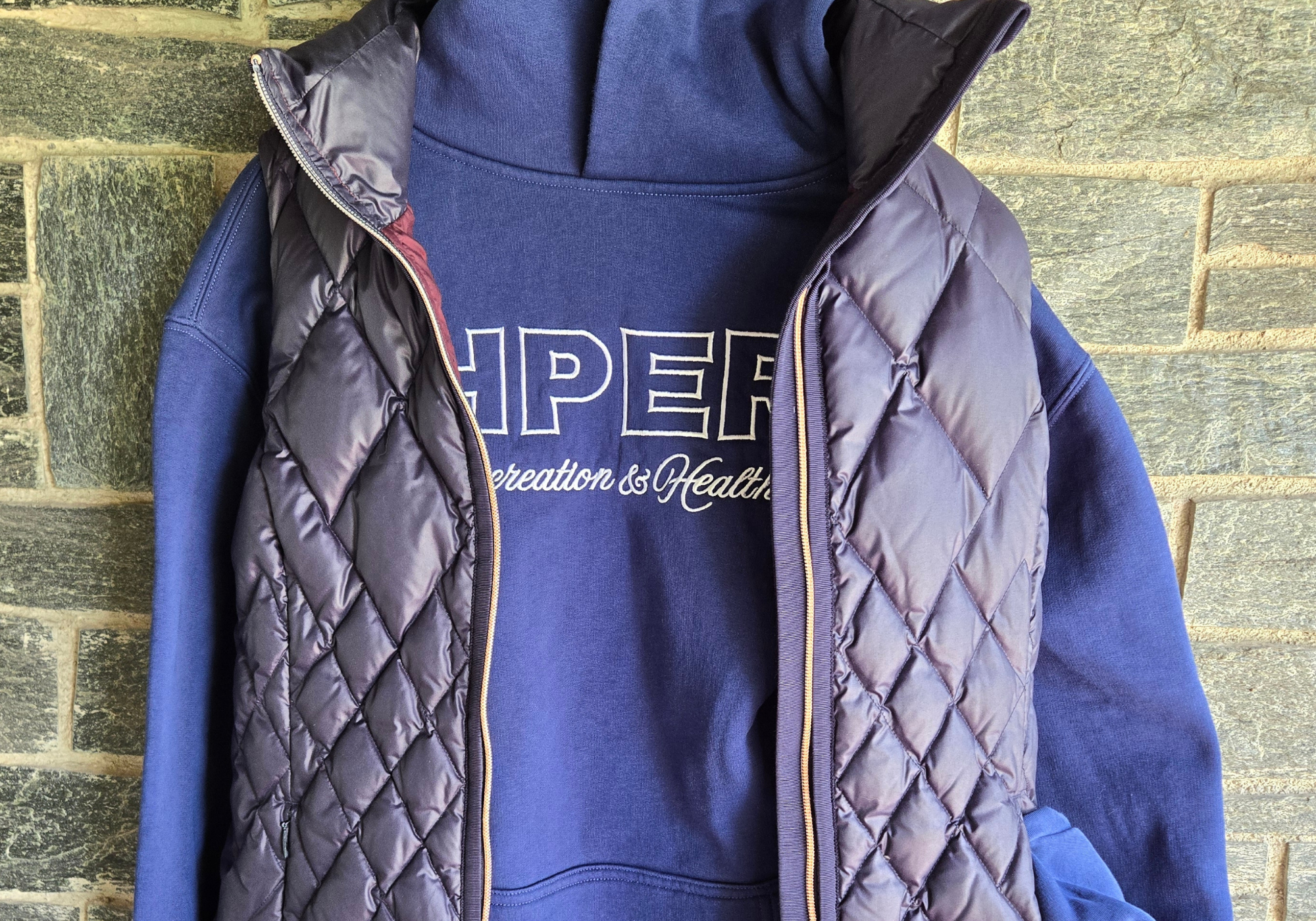 Image of a cobalt blue hoodie with a blue puffer vest hanging in front of a gray brick background.