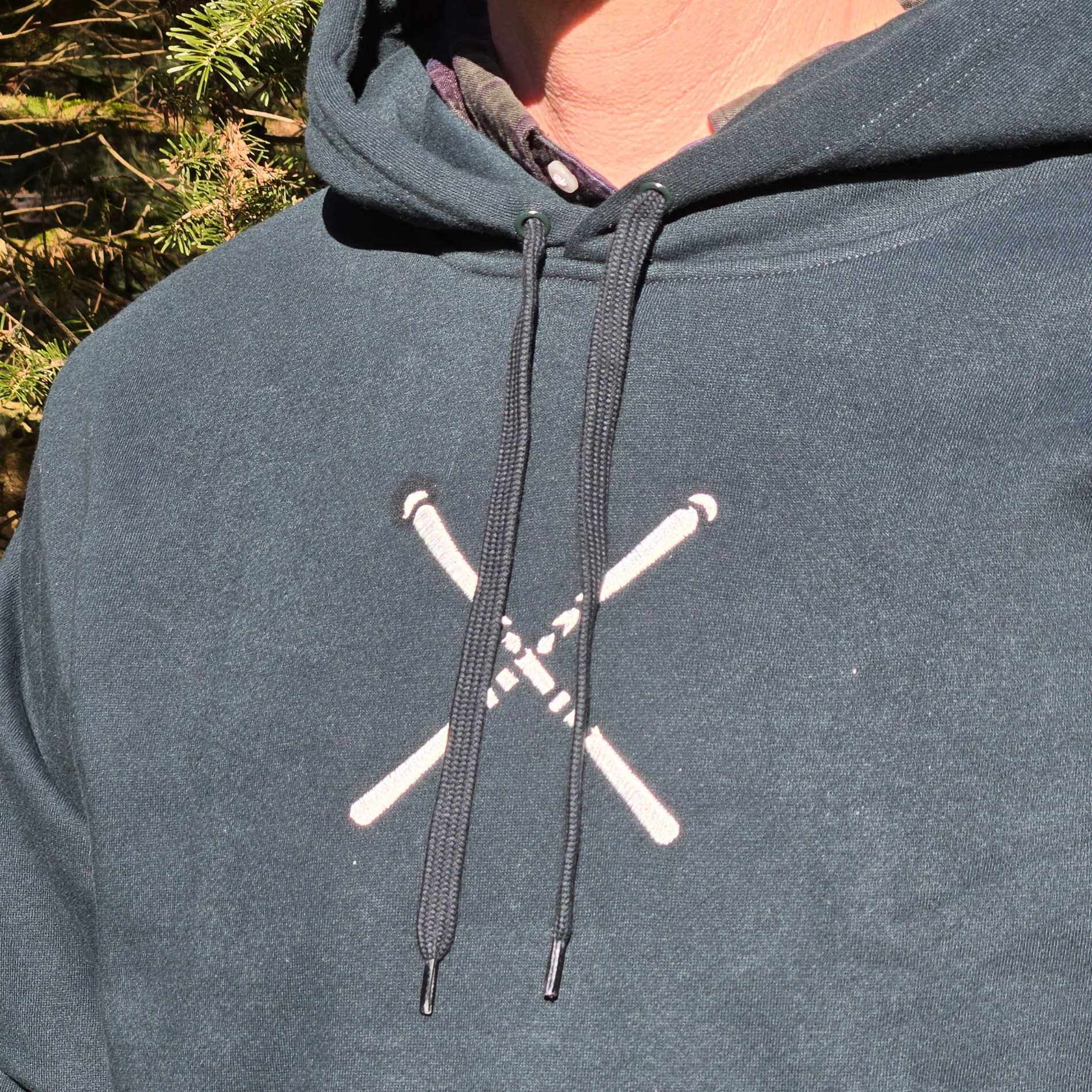 man wearing swith apres alpine club fleece hoodie in dark green with an embroidered crossed ski logo in off-white color on the center chest with drawcord visible