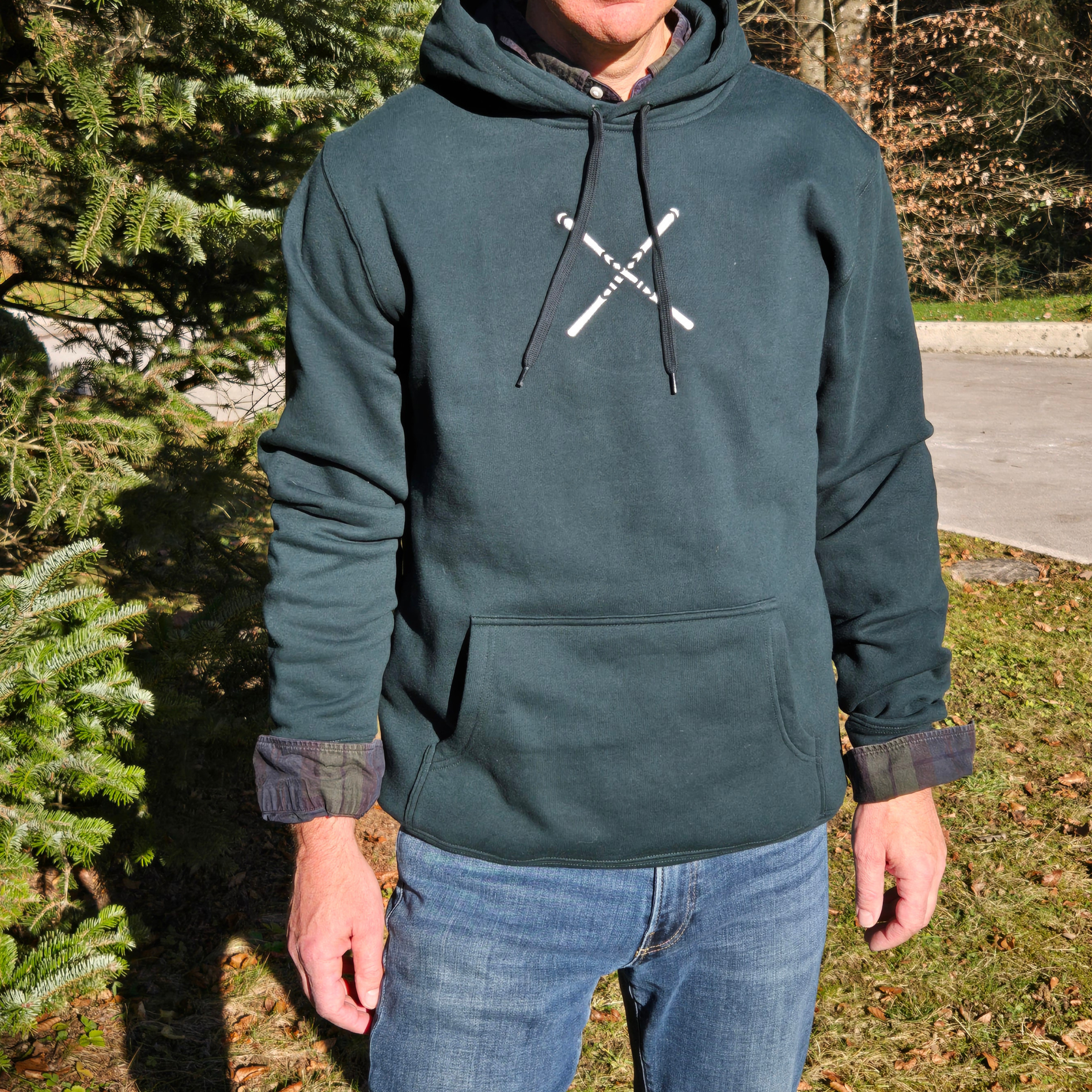 man wearing swith apres alpine club fleece hoodie in dark green with an embroidered logo of crossed skis in off-white on the center chest standing outdoors in front of a fir tree
