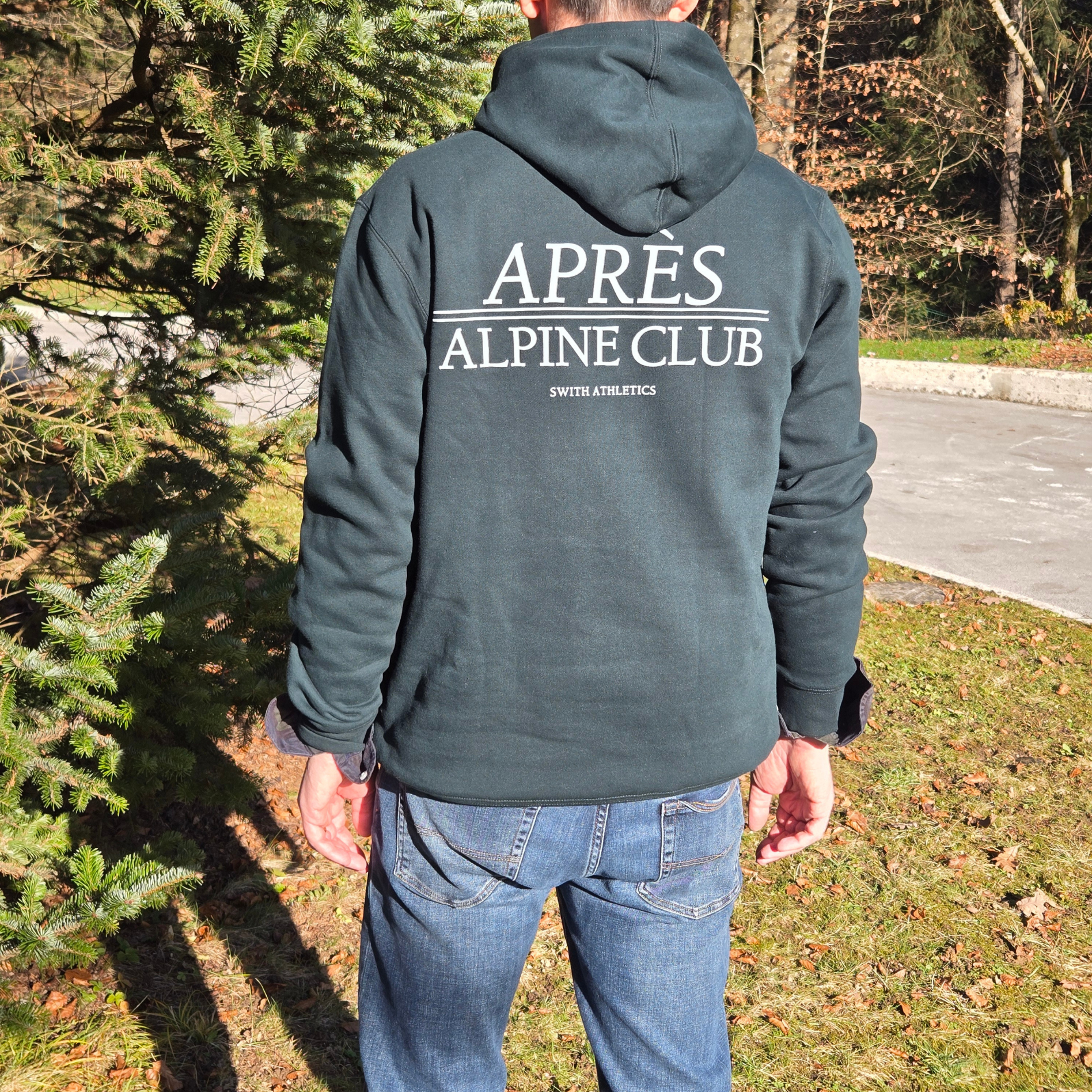 man wearing swith apres alpine club fleece hoodie with 'APRèS ALPINE CLUB' printed on the back in off-white color standing outdoors in front of a fir tree