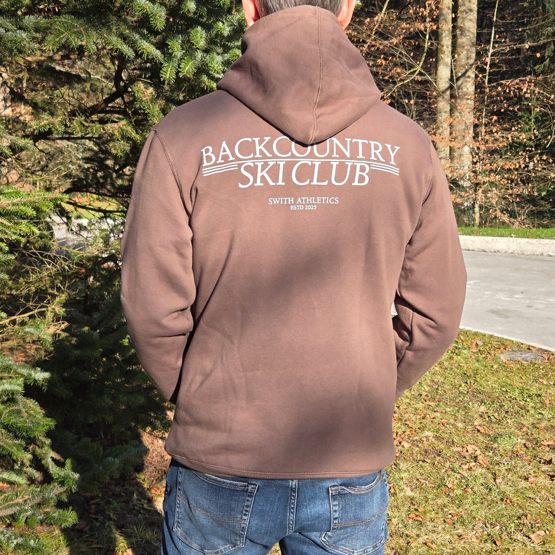 man wearing swith backcountry ski club fleece hoodie in brown with 'Backcountry Ski Club' text in off-white, standing outdoors.