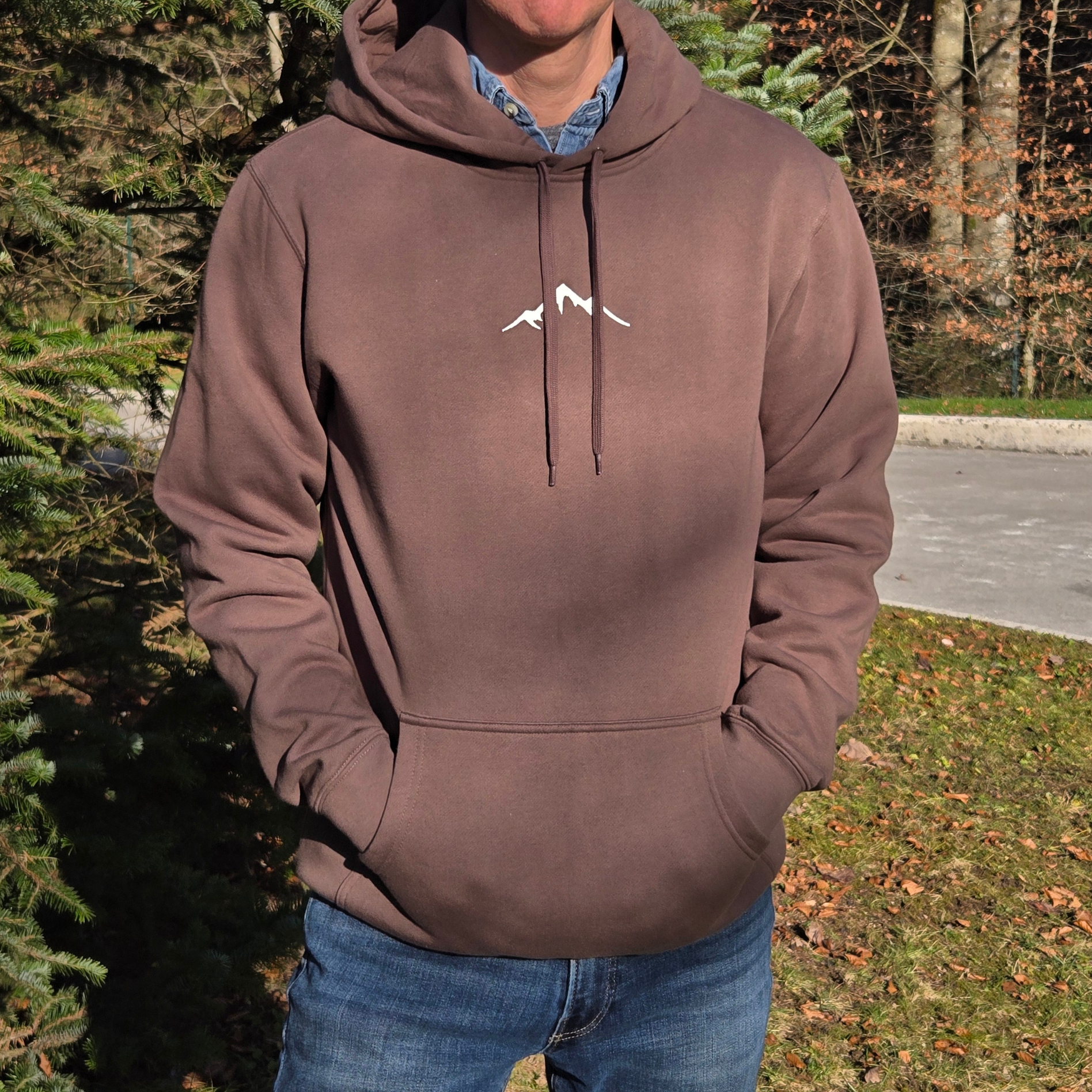 man wearing swith backcountry ski club fleece hoodie in brown with embroidered mountain outline on center chest in off-white and standing outdoors with hands in kangaroo pocket