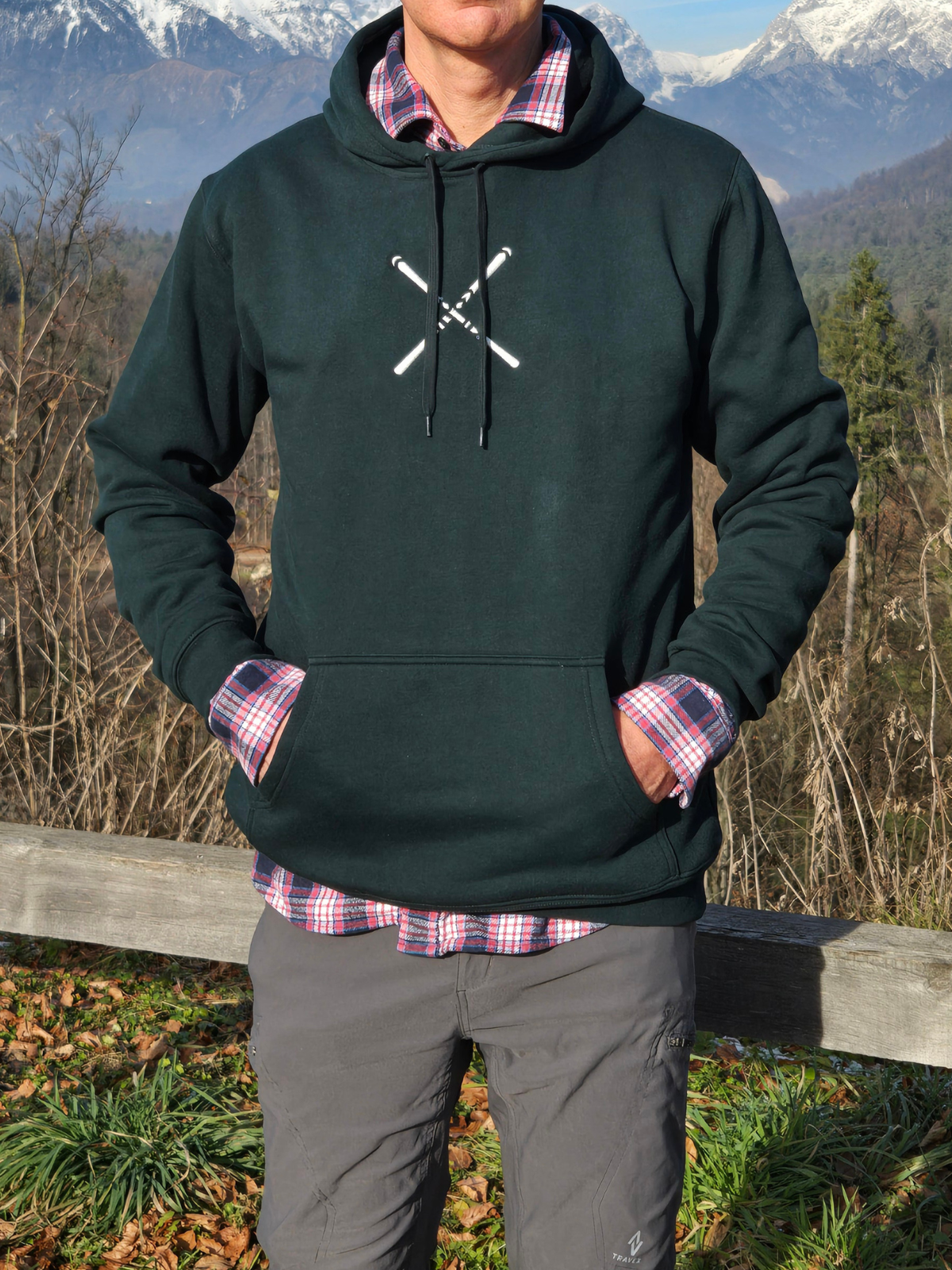 man wearing swith 'Après Alpine Club' fleece hoodie in dark green with off-white embroidery on center chest of crossed skis with snow-capped mountains in background