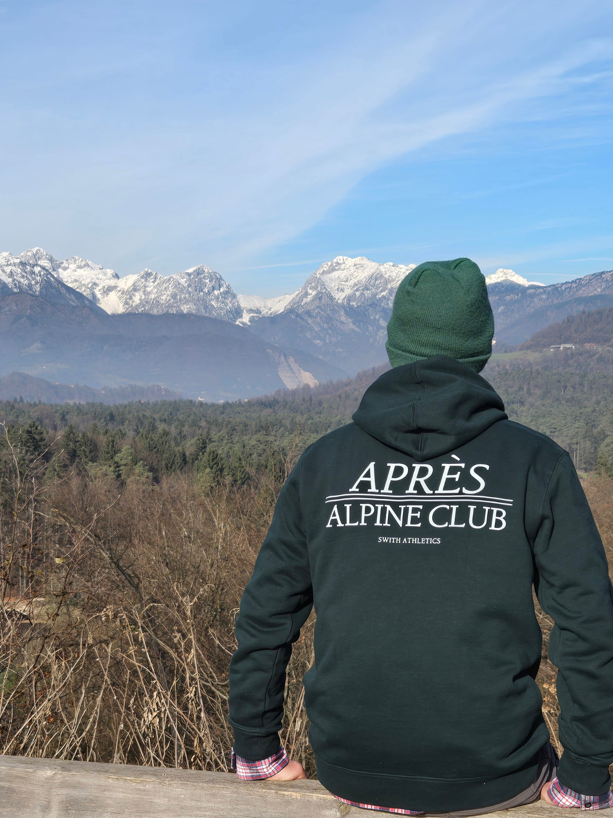 man wearing swith 'Après Alpine Club' fleece hoodie in dark green looking at snow-capped mountains.