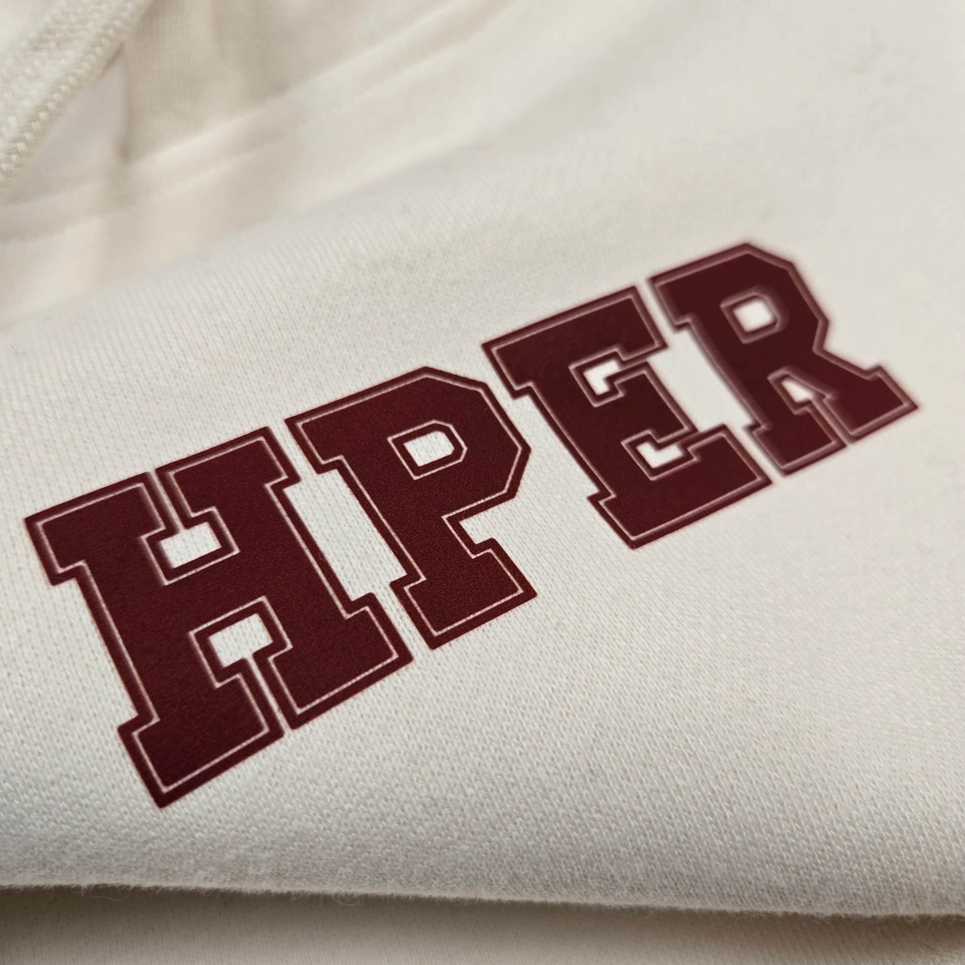 Close-up of swith hper fleece hoodie in cream color with 'HPER' text in crimson
