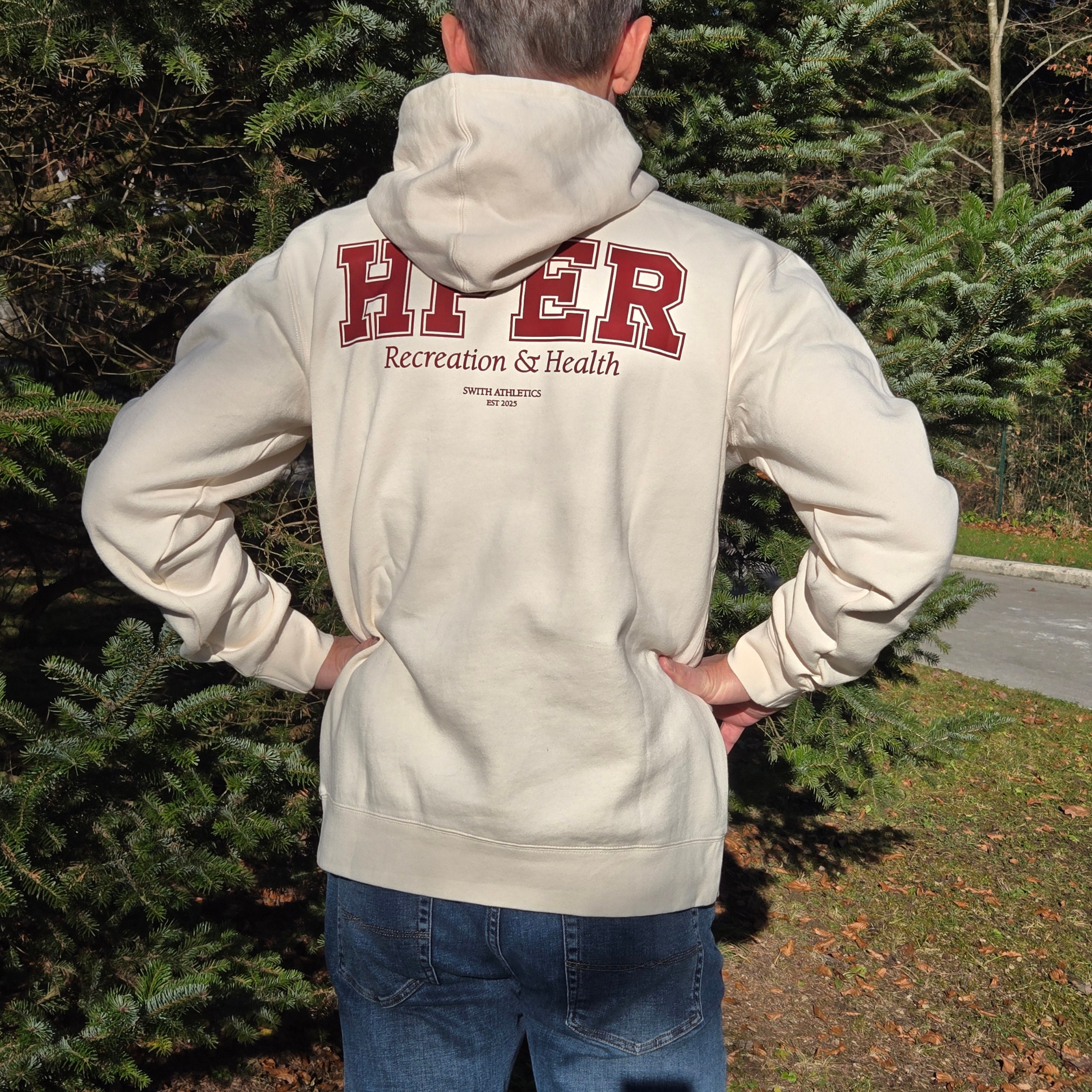 man wearing swith hper fleece hoodie in cream color with 'HPER' text on the back in crimson color, standing outdoors.