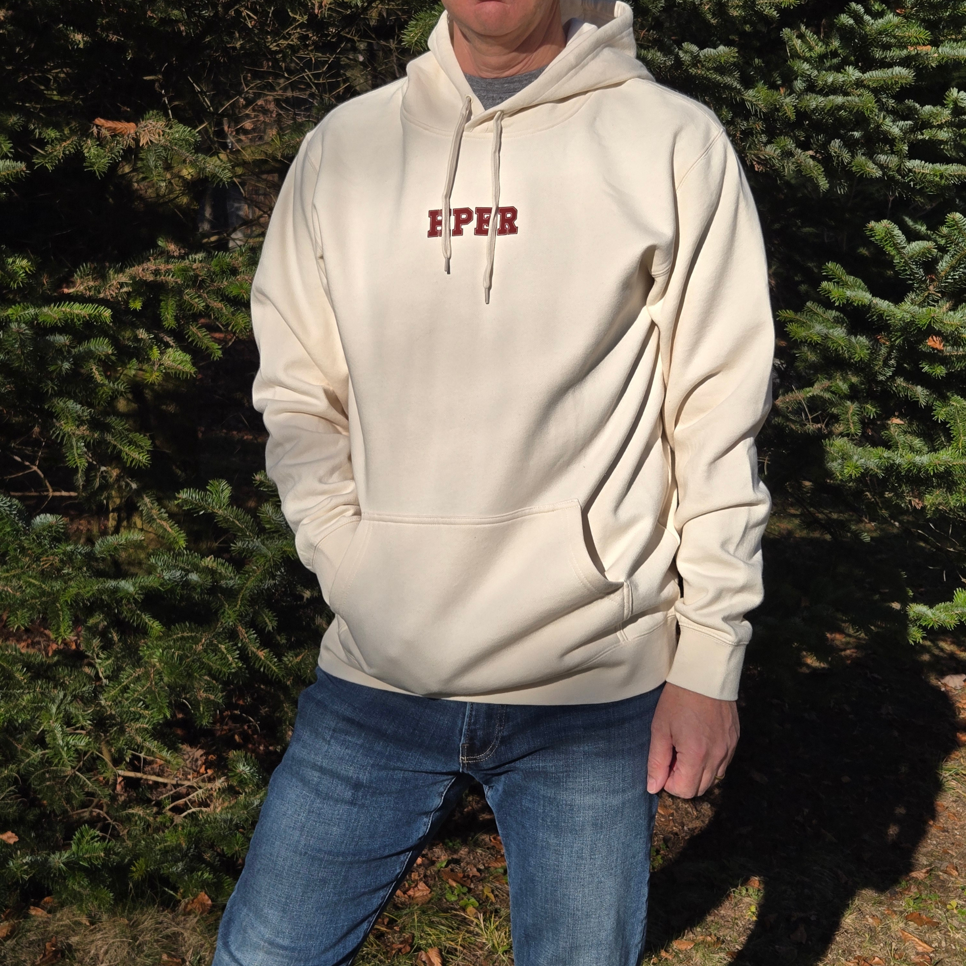 man wearing a swith hper fleece hoodie in cream color with 'hper' text in the color crimson  standing in front of trees from the front