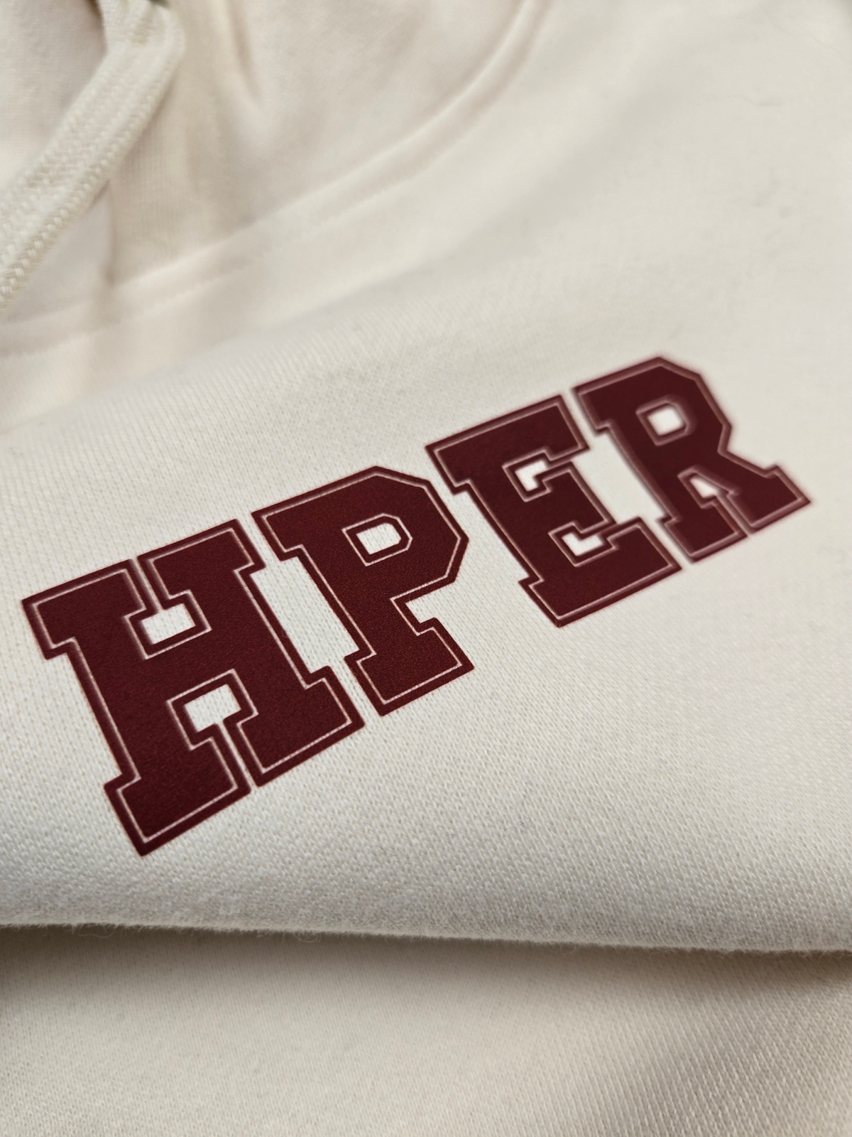 closeup of swith hper fleece hoodie with HPER text in crimson color on a cream cotton-blend fabric